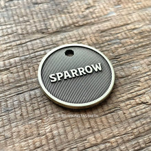 Load image into Gallery viewer, Macro product photograph of a circular, light champagne brushed brass pet tag with the name "SPARROW" in raised 3D lettering. The design features a deep fiber-laser engraved background of vertical pinstripe machined lines with a dark oxidized charcoal finish. The tag is centered on a weathered, brown horizontal-grain wooden surface with visible cracks and natural lighting.