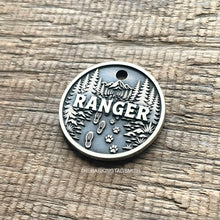 Load image into Gallery viewer, Personalised forest trail dog ID tag with engraved mountain landscape, pine trees, hiking boot prints and paw prints in brushed silver metal on rustic wood background