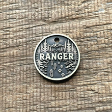 Load image into Gallery viewer, Personalised forest trail dog ID tag with engraved mountain landscape, pine trees, hiking boot prints and paw prints in brushed silver metal on rustic wood background