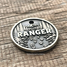 Load image into Gallery viewer, Personalised forest trail dog ID tag with engraved mountain landscape, pine trees, hiking boot prints and paw prints in brushed silver metal on rustic wood background